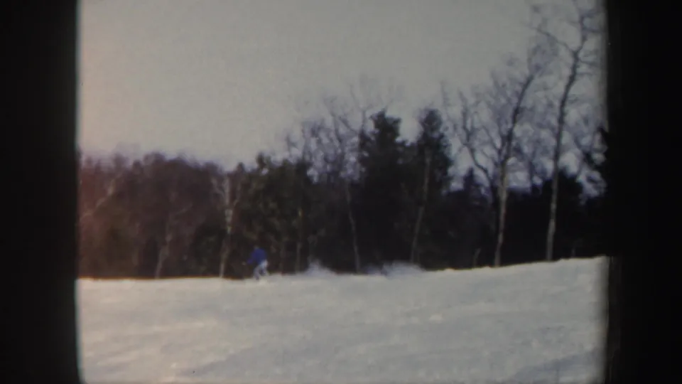 Vintage 8mm frame from Vermont showing a distant skier in blue low on the snowy slope with leafless trees behind.