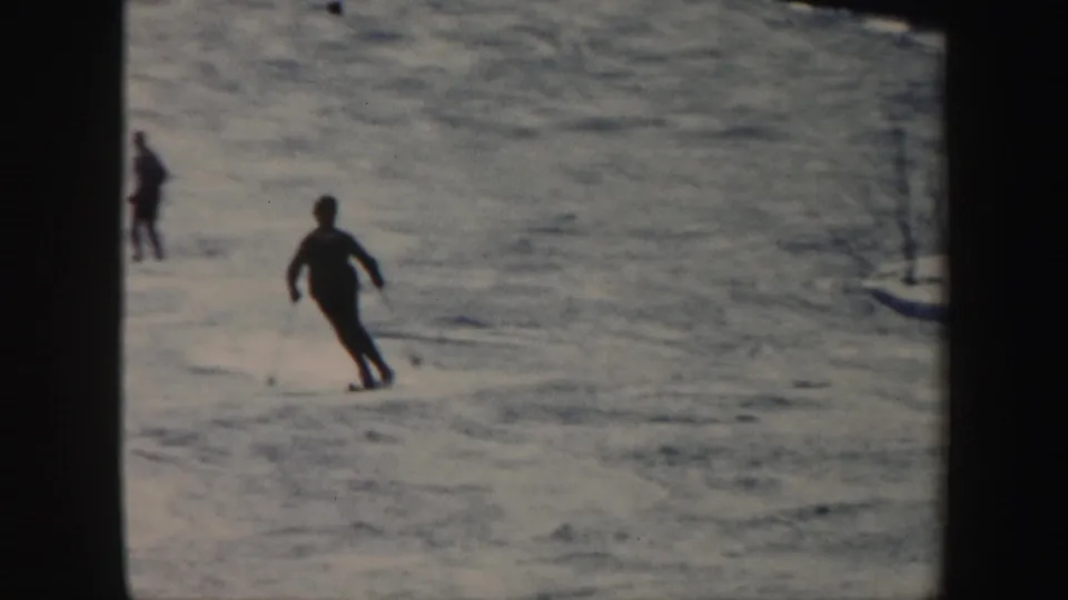 Skiers move down a Vermont snow slope, including a foreground figure turning on long skis in 1962 8mm footage.
