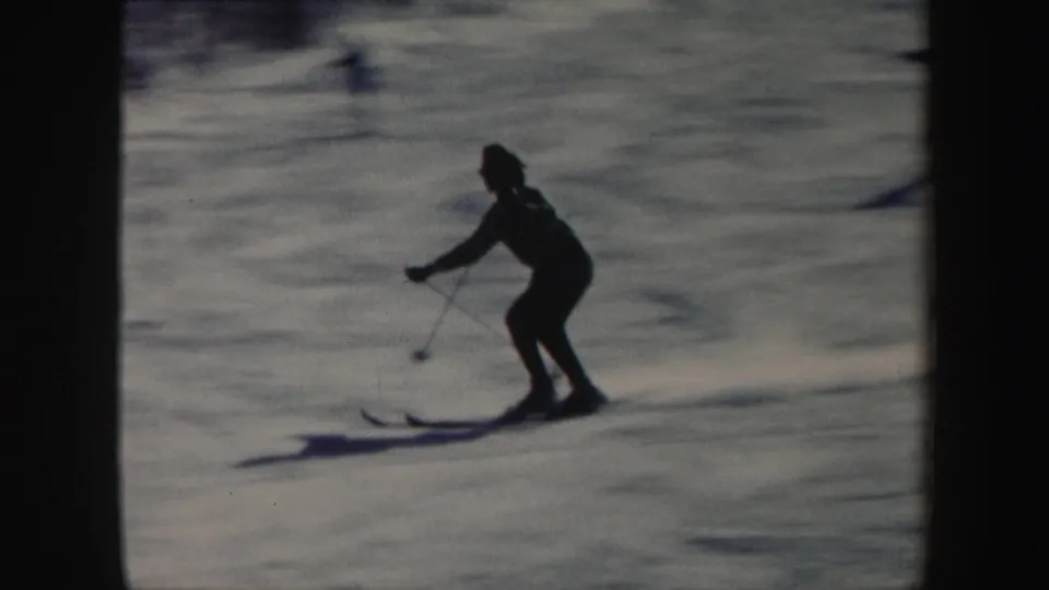 A skier in dark clothing bends forward with poles while gliding across a Vermont slope in soft 1962 home-movie footage.