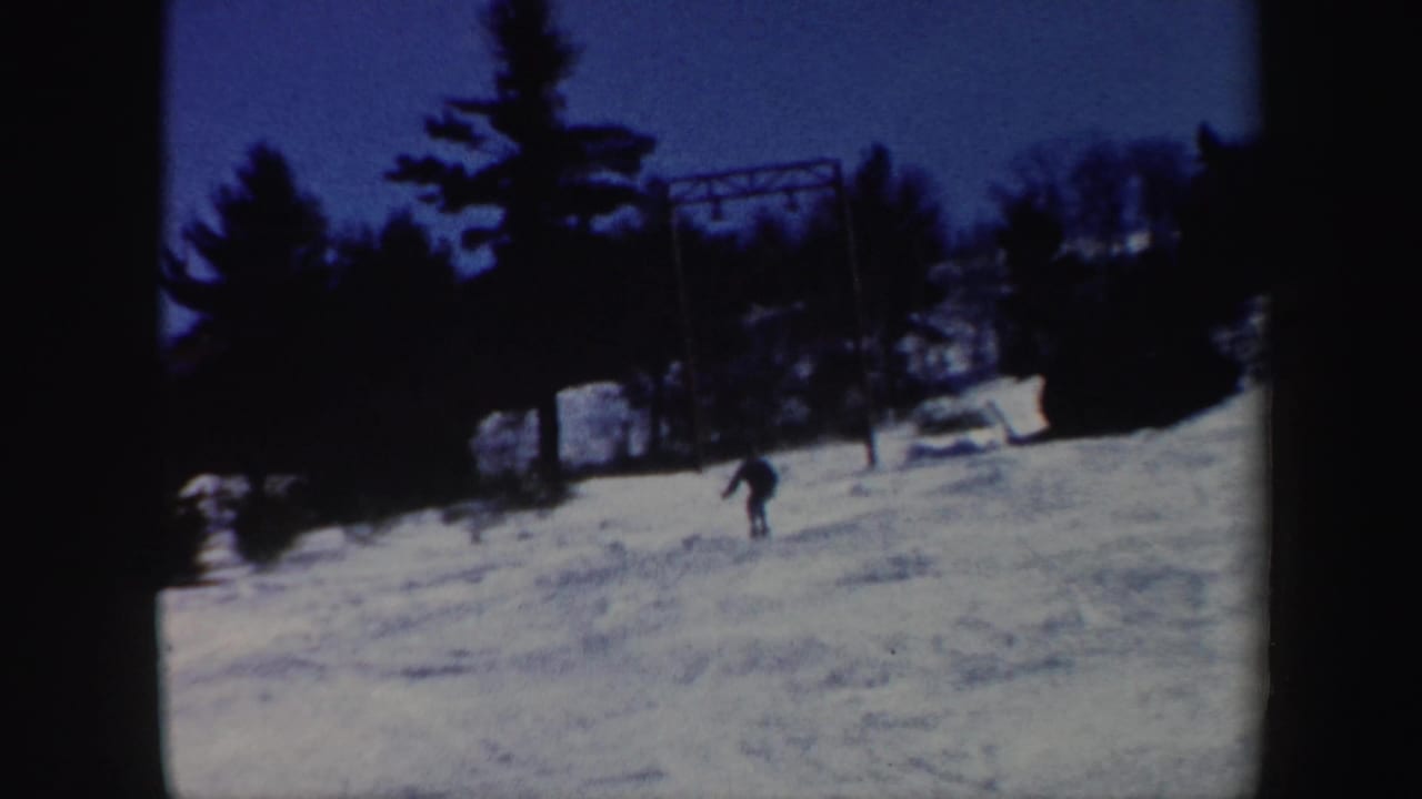 Distant skier descending a snowy slope between dark trees under a blue sky.