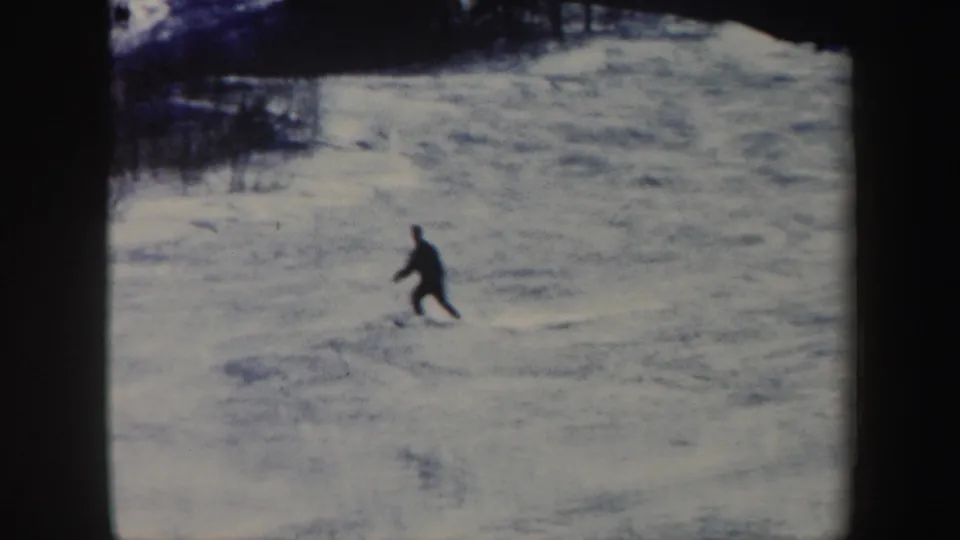 1962 Vermont home-movie frame showing a distant skier in dark clothing on a snow-covered hillside near trees.