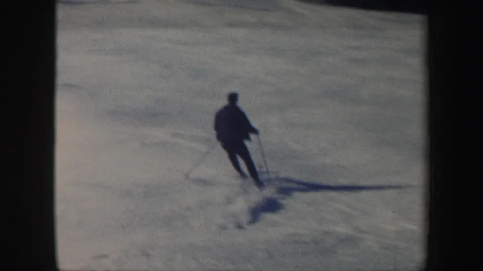 A dark-clad skier continues down the snowy Vermont hill, leaning while using poles in grainy 8mm footage.