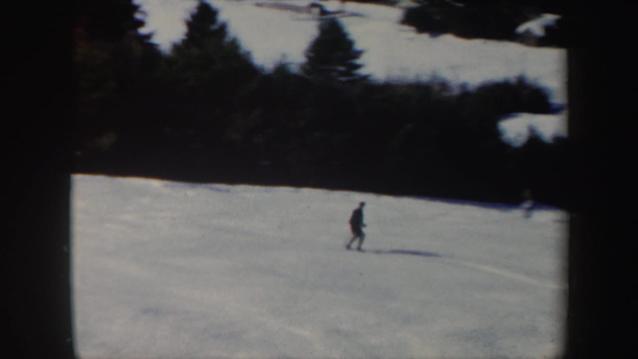 A small figure skis across a wide snowy Vermont hillside, with evergreen trees along the upper edge of the frame.
