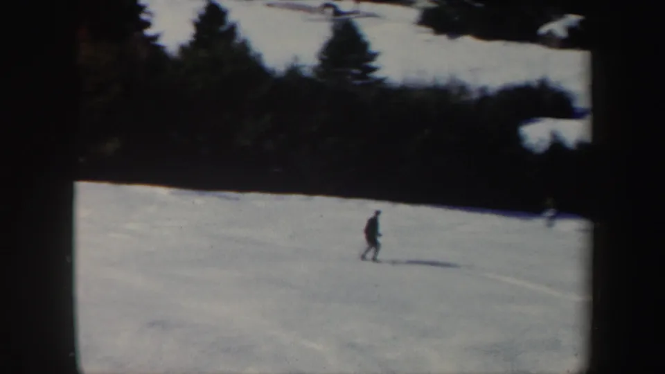 A small figure skis across a wide snowy Vermont hillside, with evergreen trees along the upper edge of the frame.