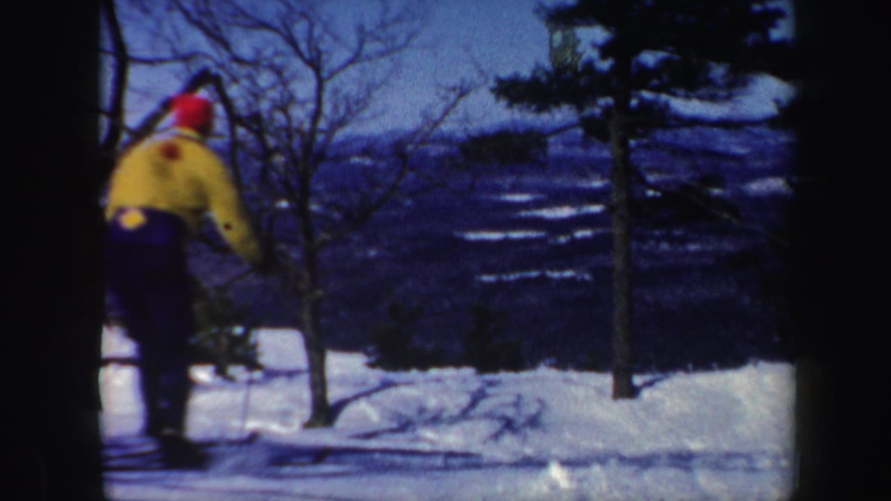 1962 Vermont home-movie frame showing a distant skier in bright winter clothing moving downhill through snow and trees.