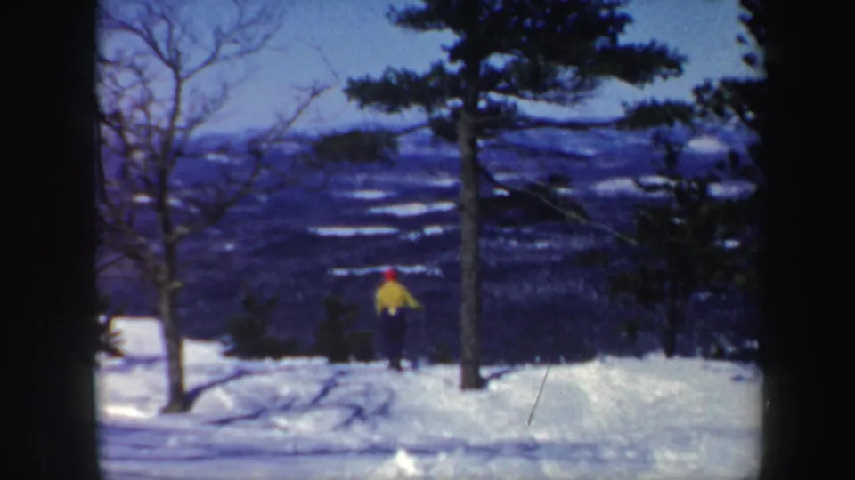The skier appears smaller in the frame while continuing downhill across a Vermont snow slope with layered mountains behind.
