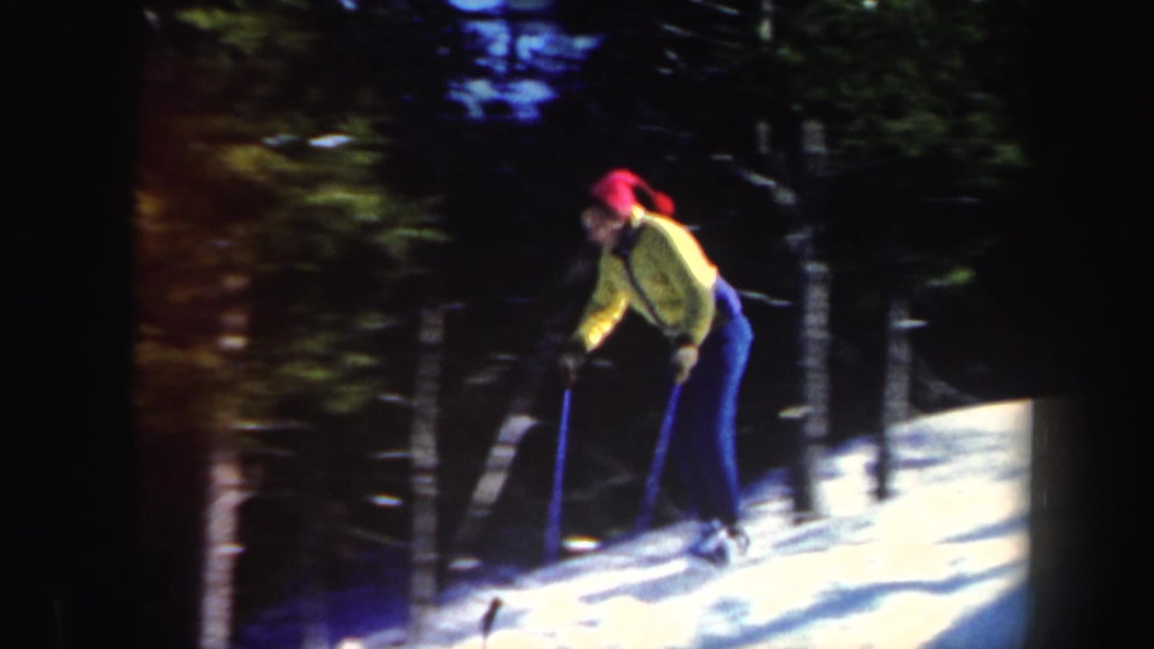 Adult skier in a yellow jacket and red cap moves across a snowy wooded slope.