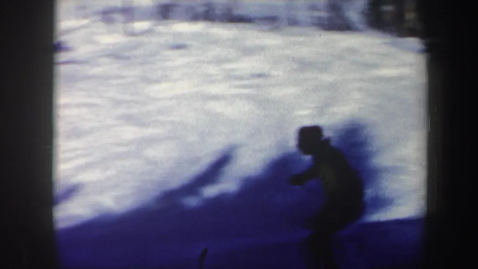 A shadowed skier passes across bright snow, with strong contrast and motion blur from the 8mm home-movie camera.