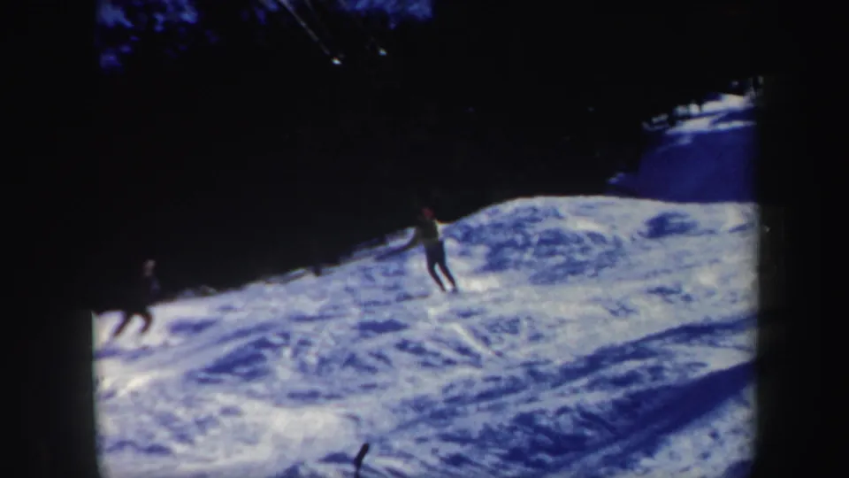 Two distant skiers appear on a snowy Vermont slope near dark woods, ending the brief 1962 home-movie skiing sequence.