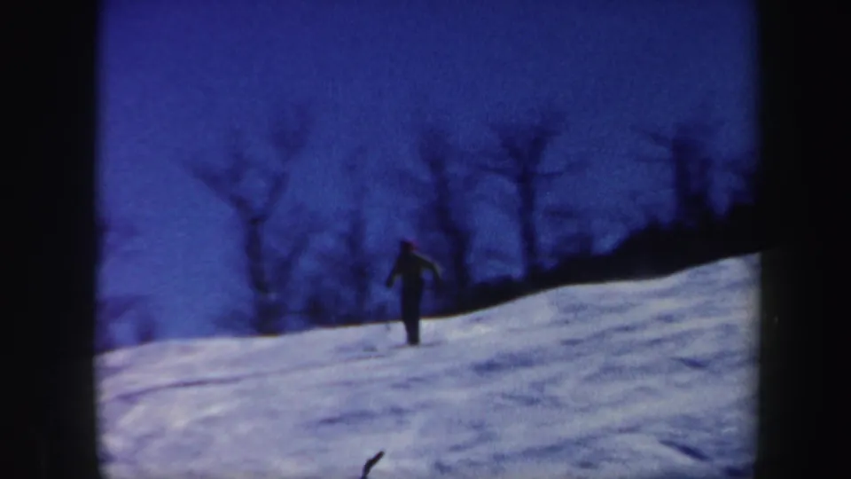 A lone skier descends the Vermont snow slope in blue-toned 1962 8mm footage, with bare trees behind them.