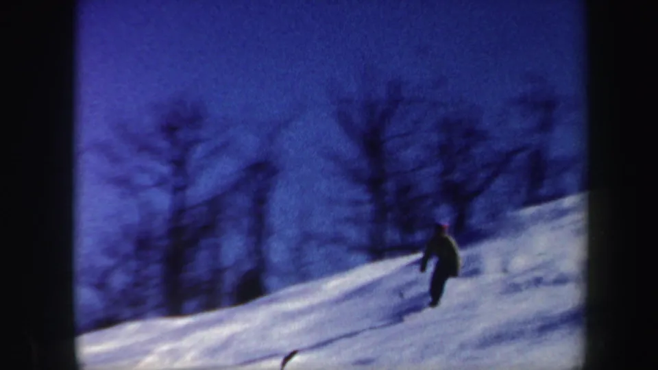In 1962 Vermont home-movie footage, a distant skier moves downhill across snow with bare trees silhouetted behind.