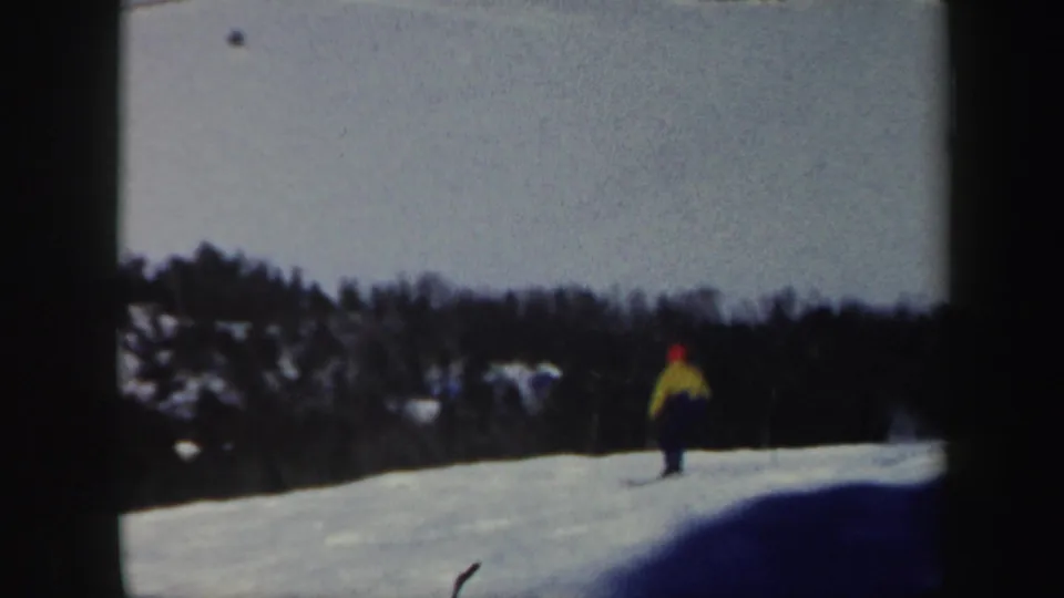 A small figure on skis descends a broad snowy slope in Vermont, framed by distant trees and dark 8mm borders.