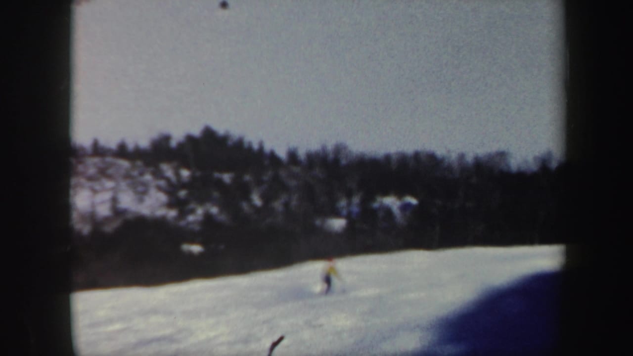 1962 Vermont home-movie frame with a skier moving across an open snowfield below a wooded ridge.
