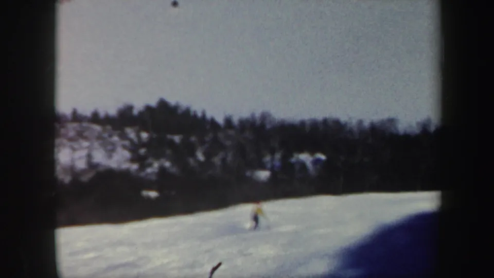 1962 Vermont home-movie frame with a skier moving across an open snowfield below a wooded ridge.