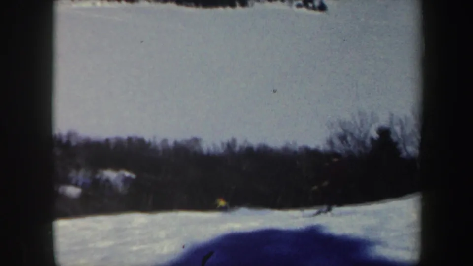1962 Vermont home-movie frame with the skier near the edge of the snowy run, partly distant against trees and shadowed snow.