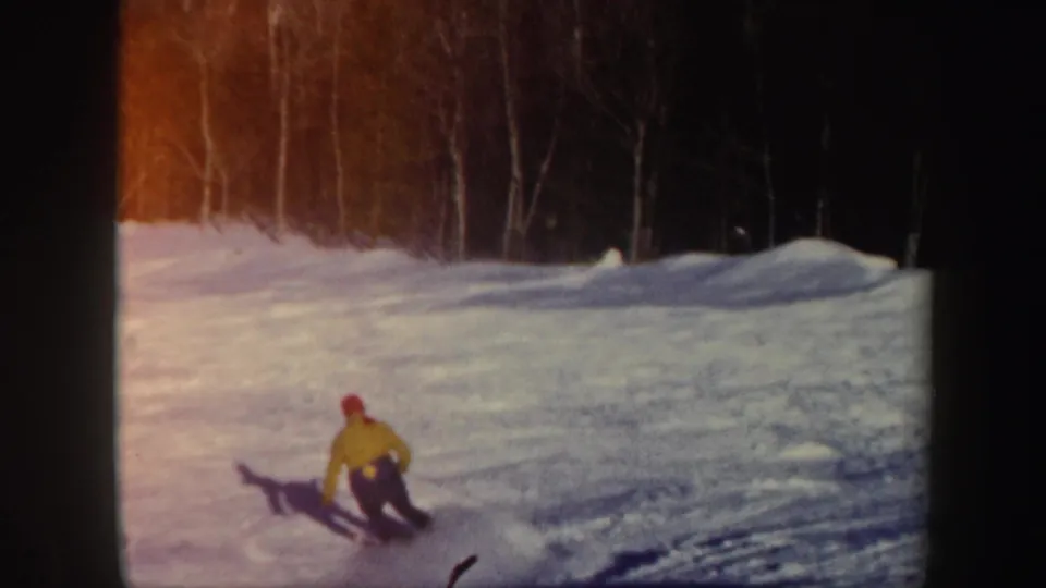 1962 Vermont 8mm frame showing a skier in a yellow jacket and red hat carving across a snowy slope near woods.