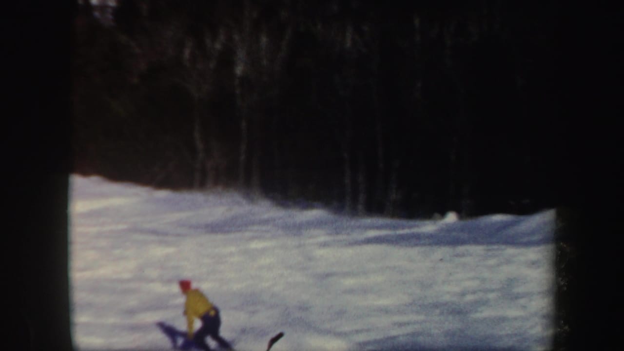 Skier in a yellow jacket and red hat moves across a snowy slope near a dark tree line.