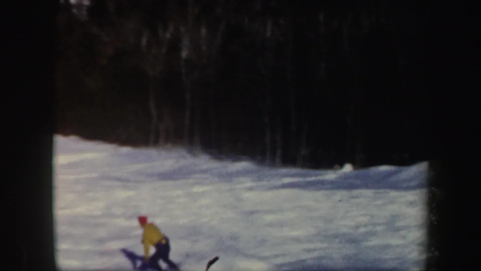 In 1962 Vermont home-movie footage, a skier in a yellow jacket and red hat descends a snowy slope bordered by trees.