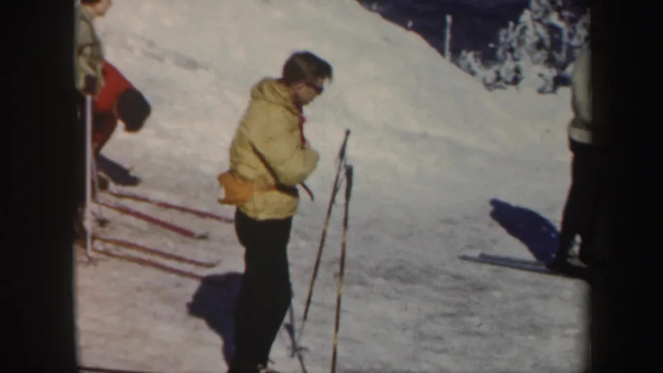 1962 Vermont ski-slope frame showing a skier in a yellow jacket adjusting gear while other skiers stand nearby.