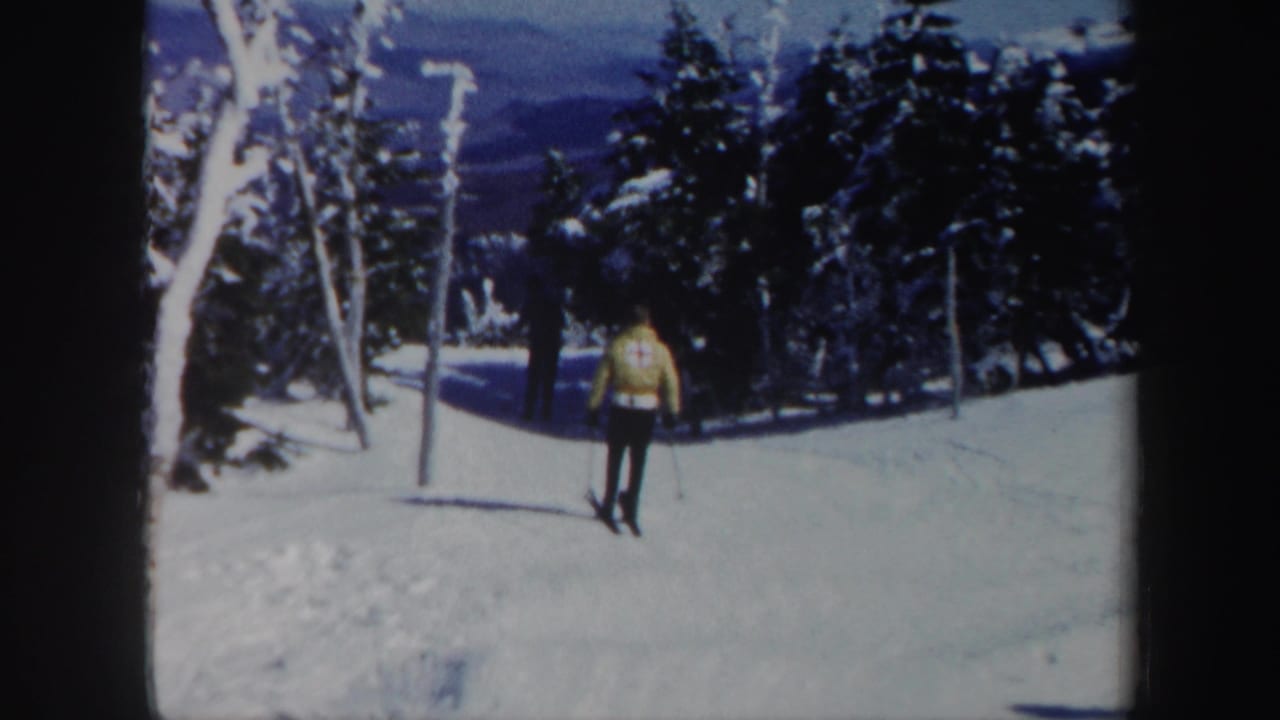 In Vermont 1962 home-movie footage, a skier in a yellow jacket moves on a snow-covered slope with evergreen trees and distant mountains behind.
