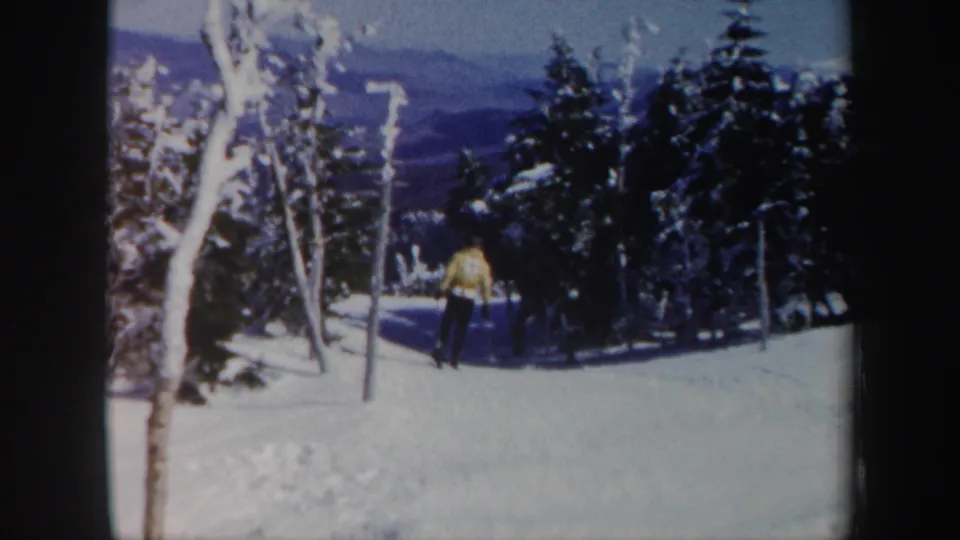 A skier in a yellow jacket moves toward a shadowed tree line on the Vermont slope, with snow, evergreens, and mountains visible in the 8mm frame.