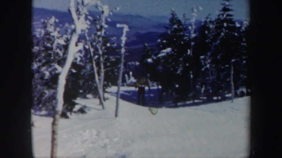 Near the end of the Vermont 1962 clip, the skier continues away along the wooded snowy slope, with mountains faintly visible beyond the trees.