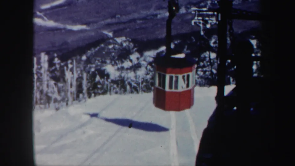 1962 Vermont 8mm frame showing a red lift cabin or gondola near a snowy ski slope, with dark lift structure and mountain forest behind it.