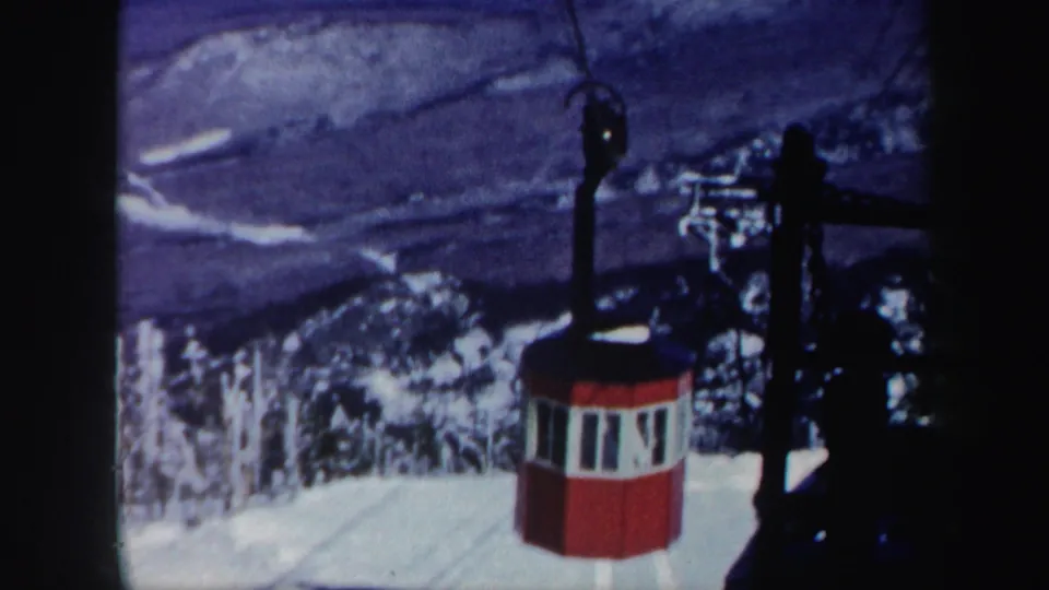 1962 Vermont ski resort frame with a red gondola-like lift cabin, dark support hardware, snowfields, trees, and mountain ridges beyond.