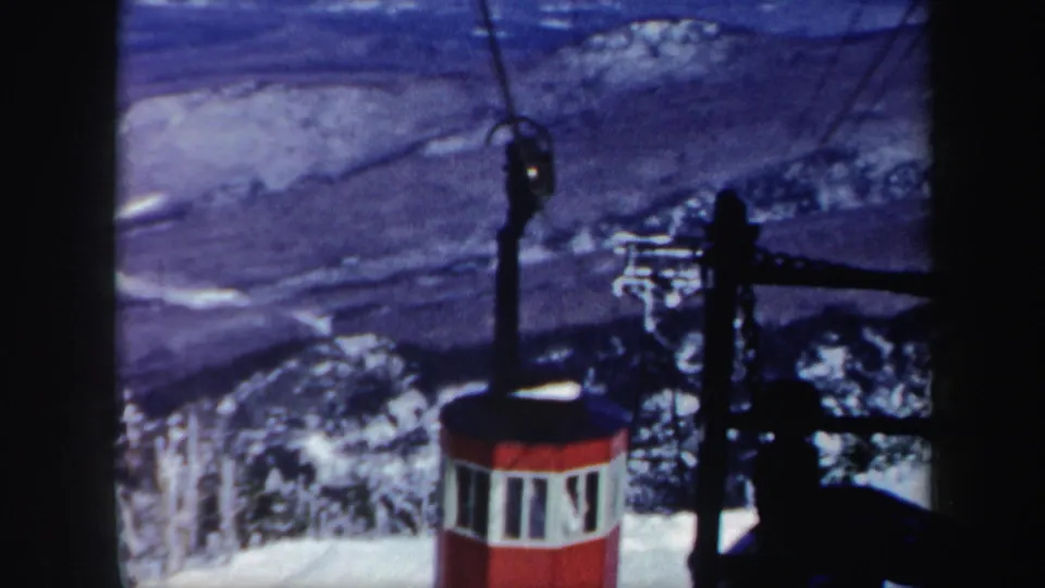 1962 Vermont home-movie frame showing a red cable car with white windows moving above snow-covered slopes and trees.