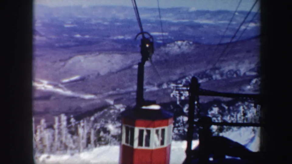 A red cable car travels along overhead wires above snow-covered trees and mountain terrain in the Vermont winter footage.