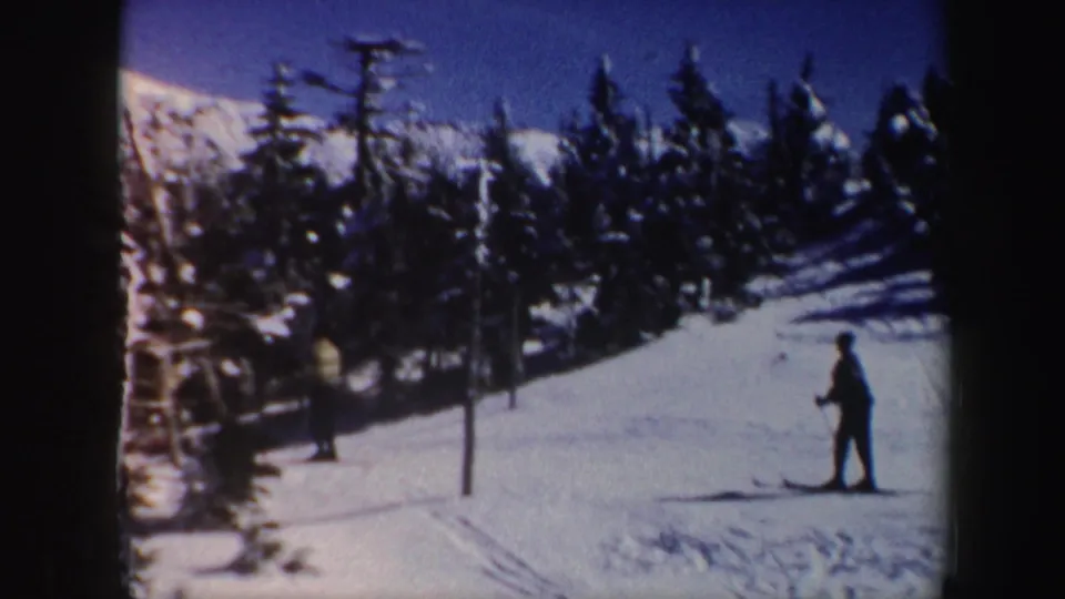 A lone skier stands or glides on a Vermont ski trail, with snow-covered evergreens and lift poles behind.