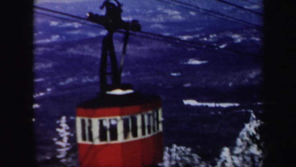 A red mountain cable car rides below overhead cables, seen against dark snowy slopes in 1962 Vermont footage.