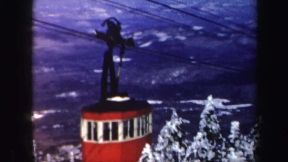 The red cable car continues along overhead wires above snow-covered evergreens and mountain terrain.