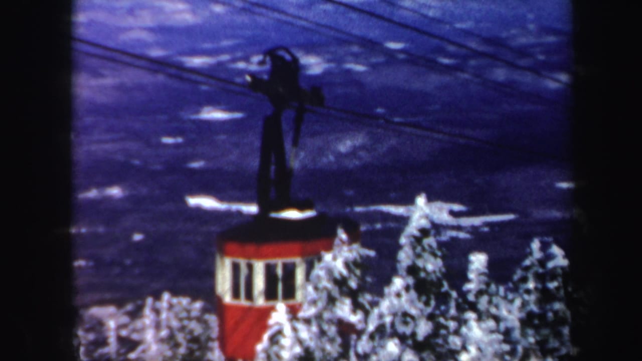 1962 Vermont 8mm frame showing a red cable car on overhead lines above snow-laden conifers and a blue winter mountain backdrop.