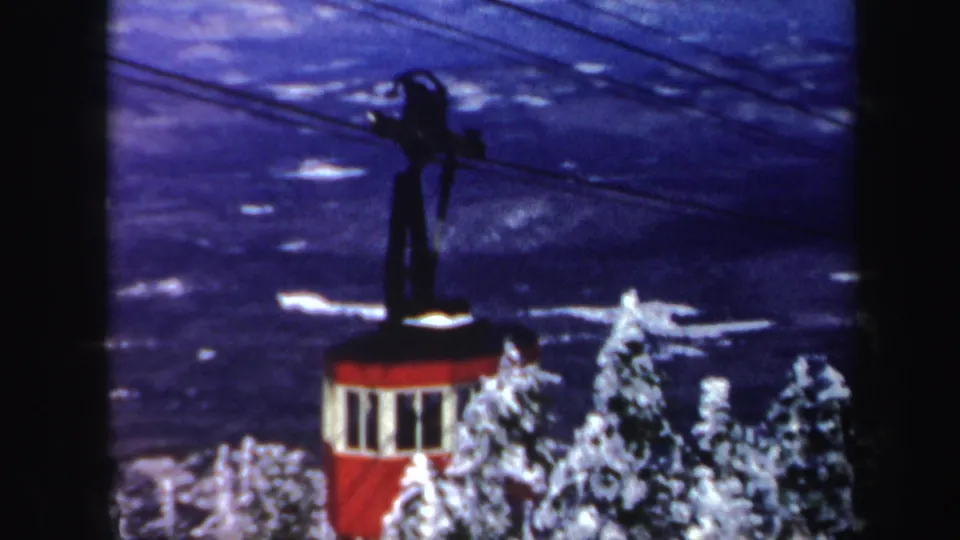 1962 Vermont 8mm frame showing a red cable car on overhead lines above snow-laden conifers and a blue winter mountain backdrop.