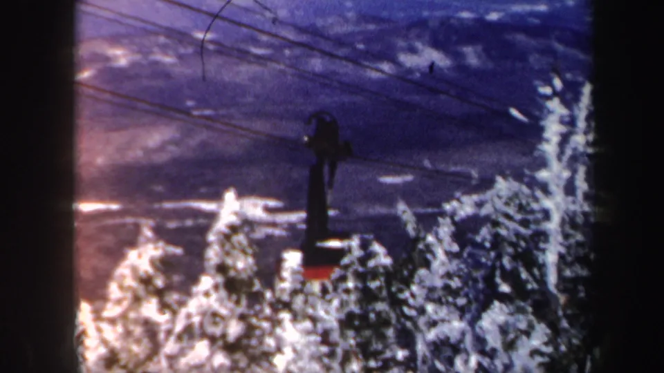 1962 Vermont 8mm frame showing the cable car hardware silhouetted over dense snow-laden conifers in a ski-area landscape.