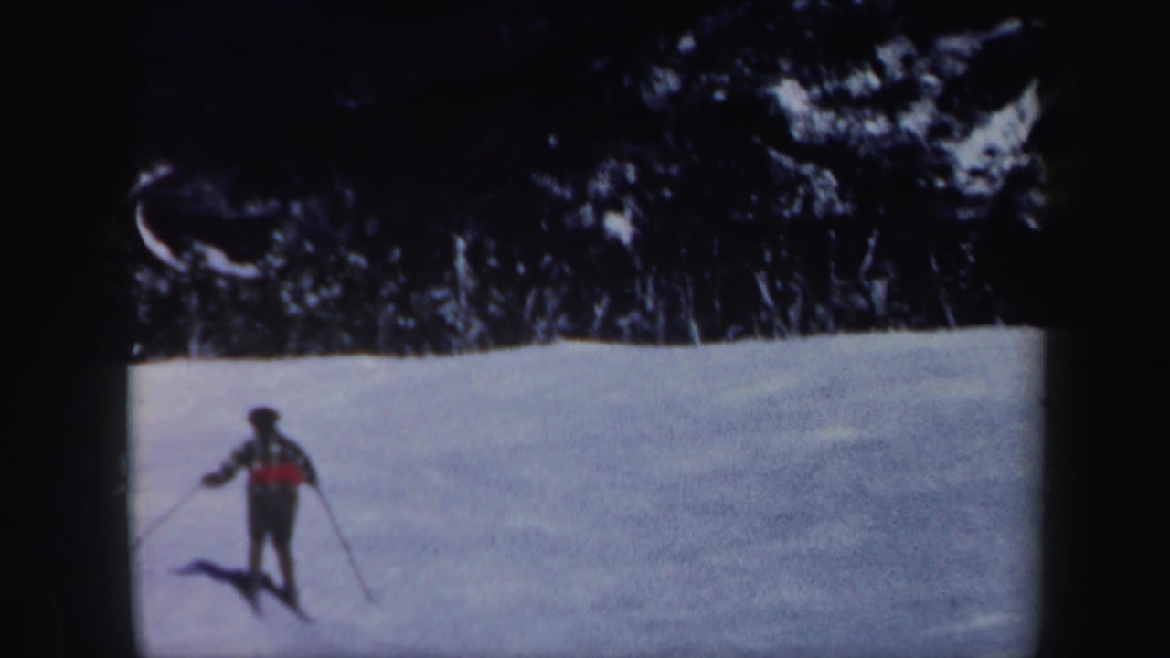 Skier in a red plaid jacket standing or moving on a snowy slope near a dark treeline.