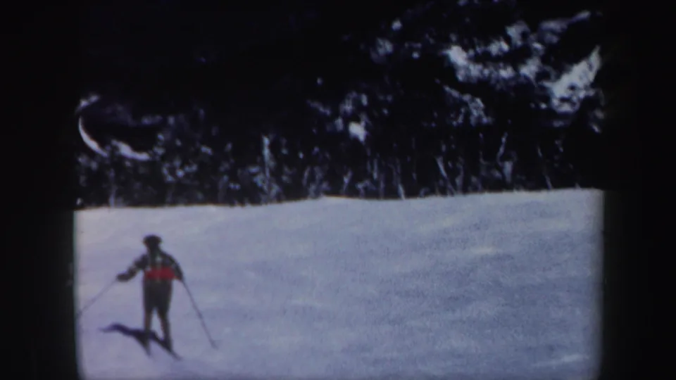 1962 Vermont 8mm frame of a skier in a red plaid jacket on a bright snowy slope below dark winter trees.