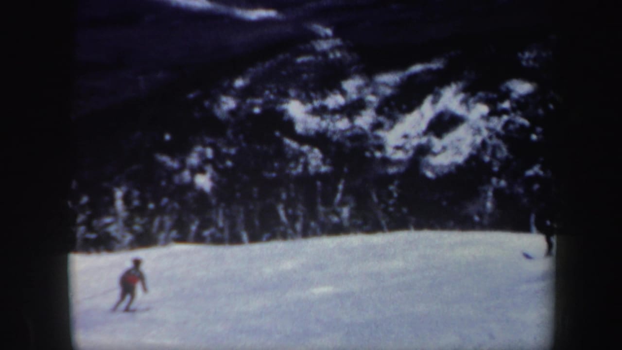 1962 Vermont home-movie frame showing a distant skier moving across an open snowy slope beneath a dark treeline.