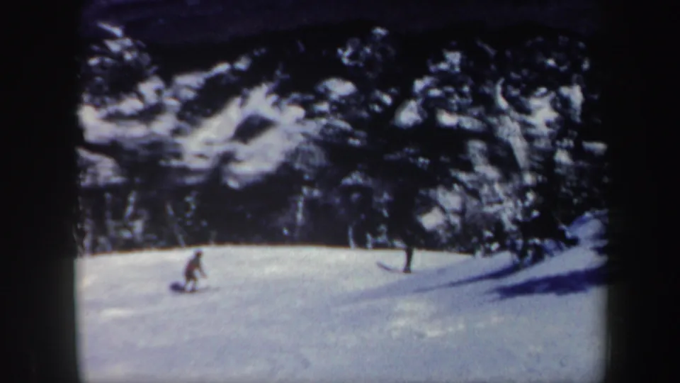 1962 Vermont 8mm frame of a small skier descending a white slope bordered by snow-dusted trees.