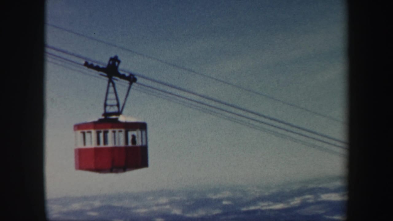 Red gondola cable car suspended above snowy mountains under a pale sky.