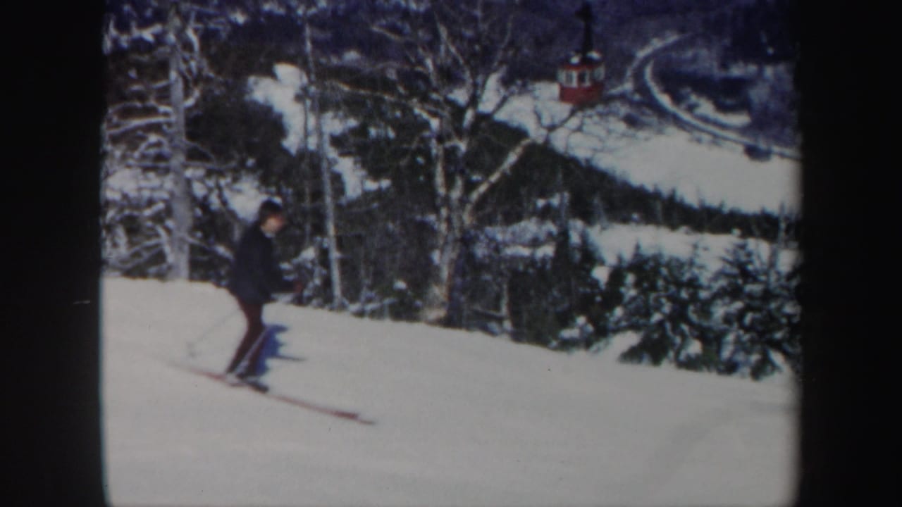 Man skiing down a snowy slope near bare trees and a distant red lift car.