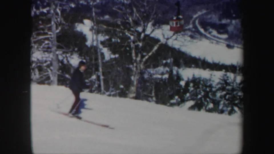 In Vermont 1962 home-movie footage, a man skis across a snowy slope with trees and a red lift car in the background.