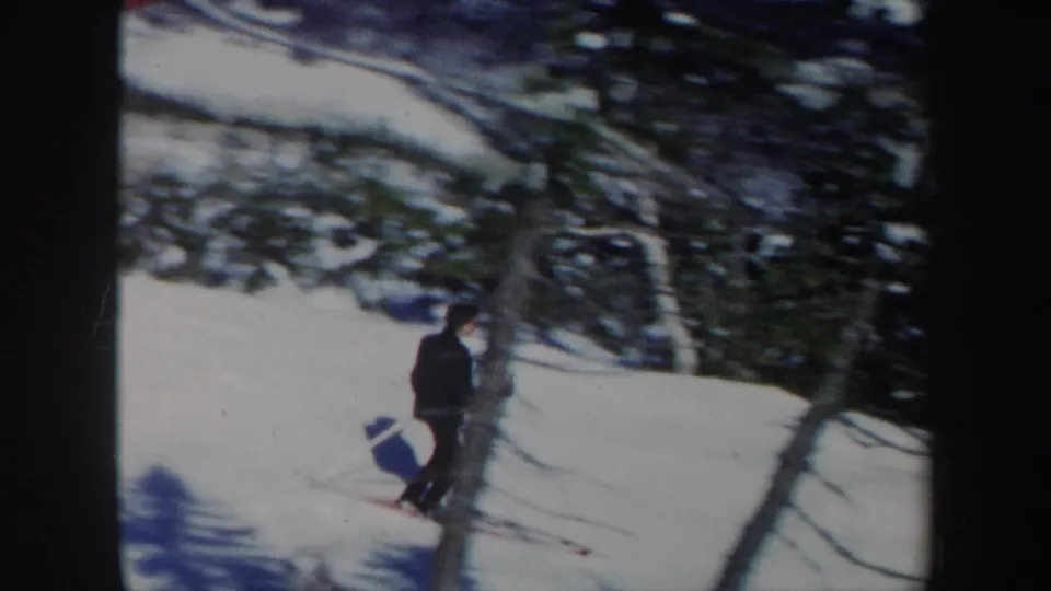 A dark-clad skier continues downhill in Vermont, with snow, trees, and distant ski-area landscape visible behind him.