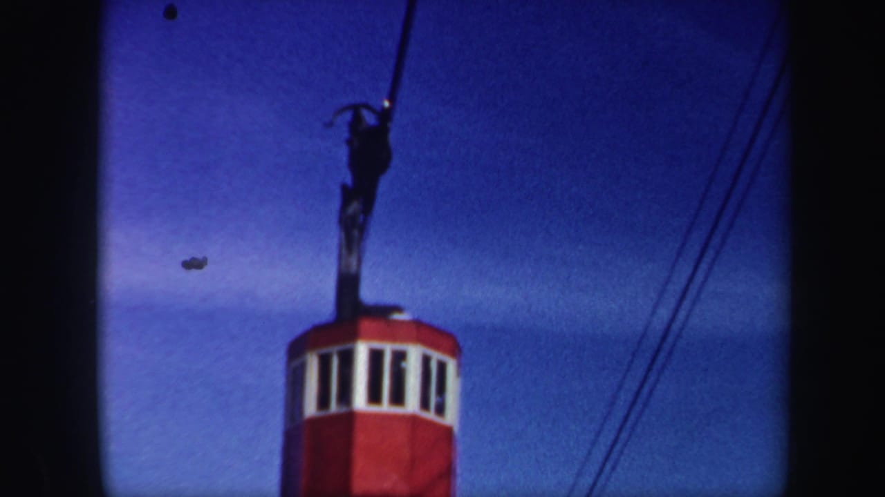 1962 Vermont 8mm frame of a red ski-lift cabin ascending beneath dark cable hardware against a saturated blue sky.