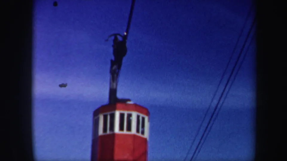 1962 Vermont 8mm frame of a red ski-lift cabin ascending beneath dark cable hardware against a saturated blue sky.