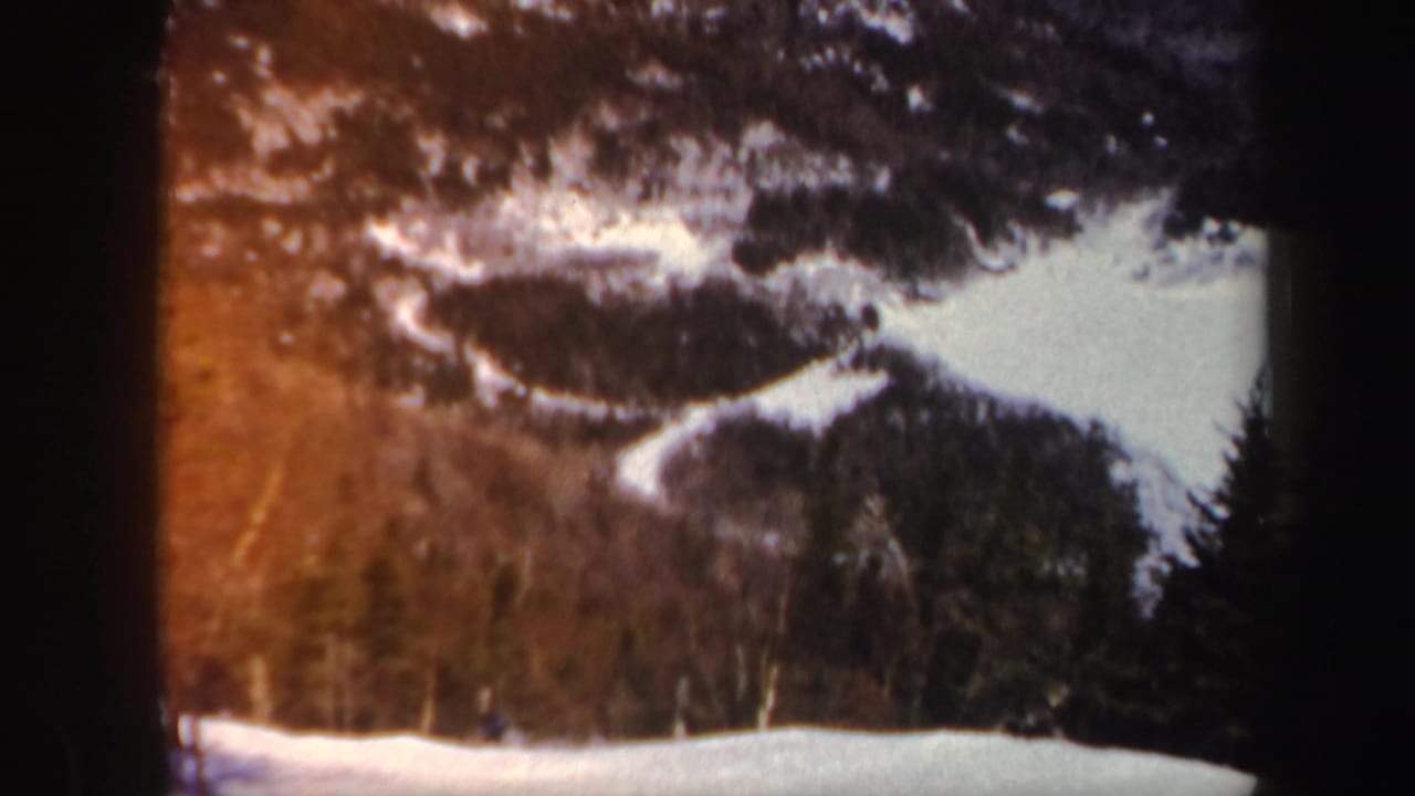 Warm-toned frame of snowy mountain terrain with trees and a small figure on the slope.
