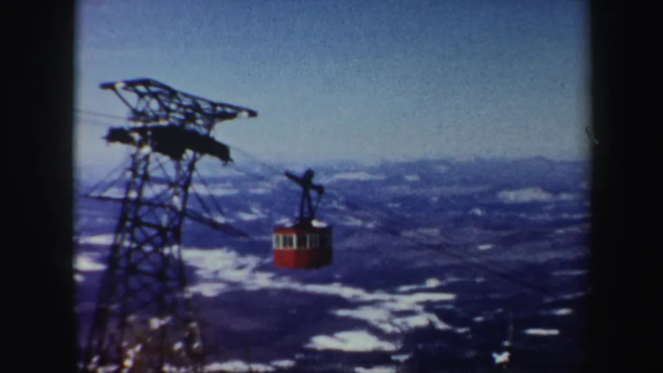 1962 Vermont 8mm frame showing a red cable car near a lattice support tower over a snow-covered mountain landscape.