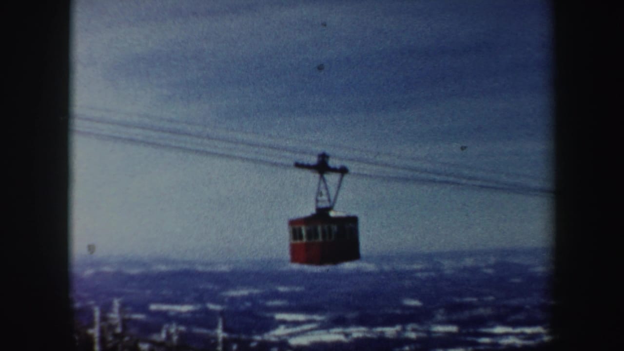 1962 Vermont home-movie frame showing a red aerial cable car crossing above snowy hills under a cloudy sky.