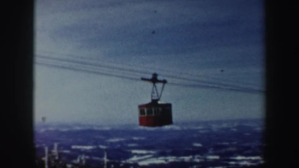 1962 Vermont home-movie frame showing a red aerial cable car crossing above snowy hills under a cloudy sky.